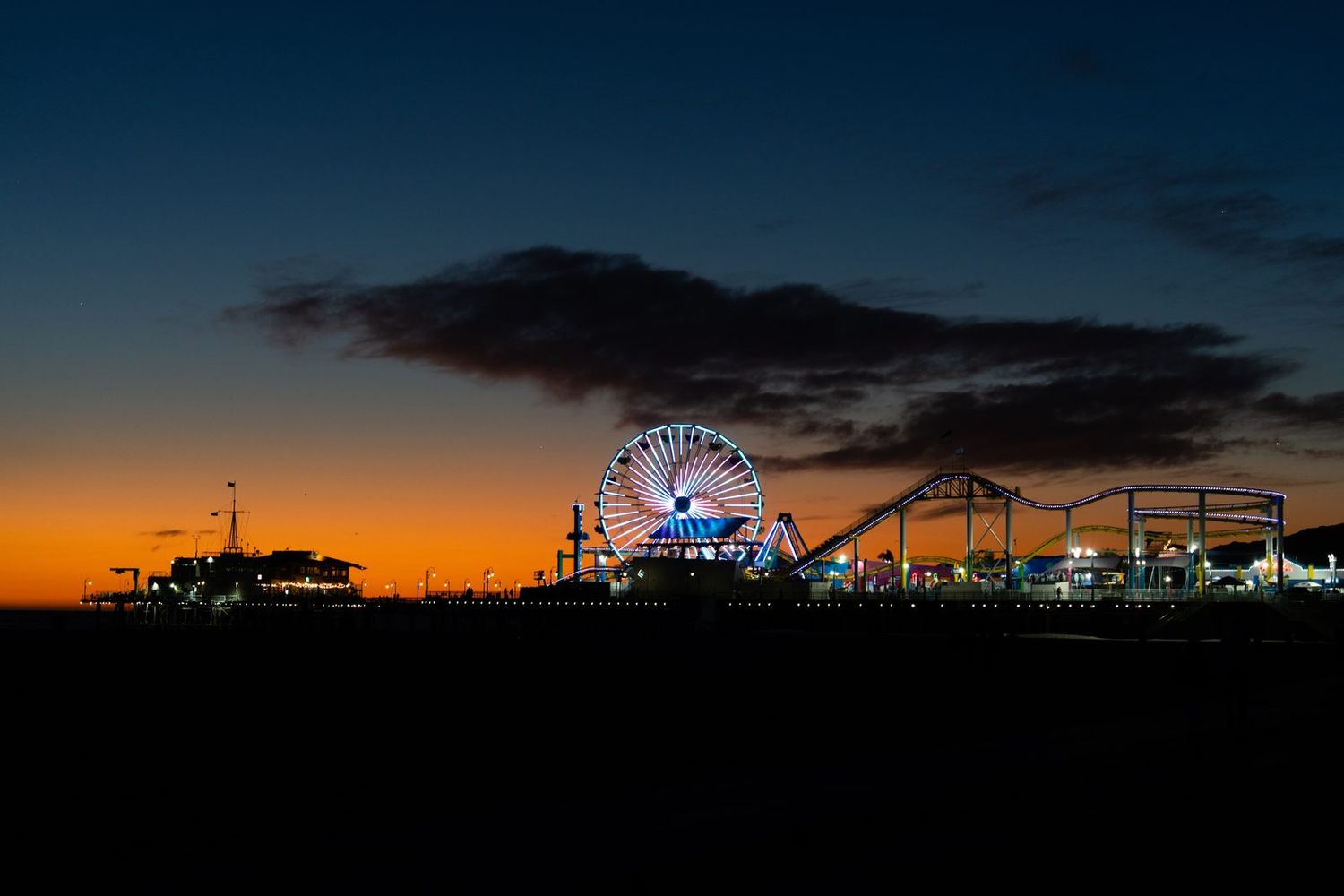 The lit Ferris wheel and roller coaster of Santa Monica Pier against a blue-hour sky